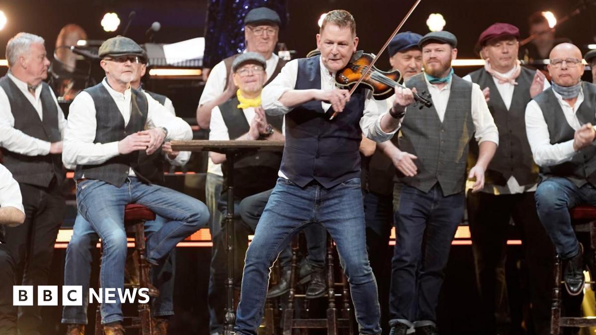Mike McGrother performs on the violin on stage backed by his choir who are standing behind him with their traditional uniform of waistcoat, white shirts and flat caps.