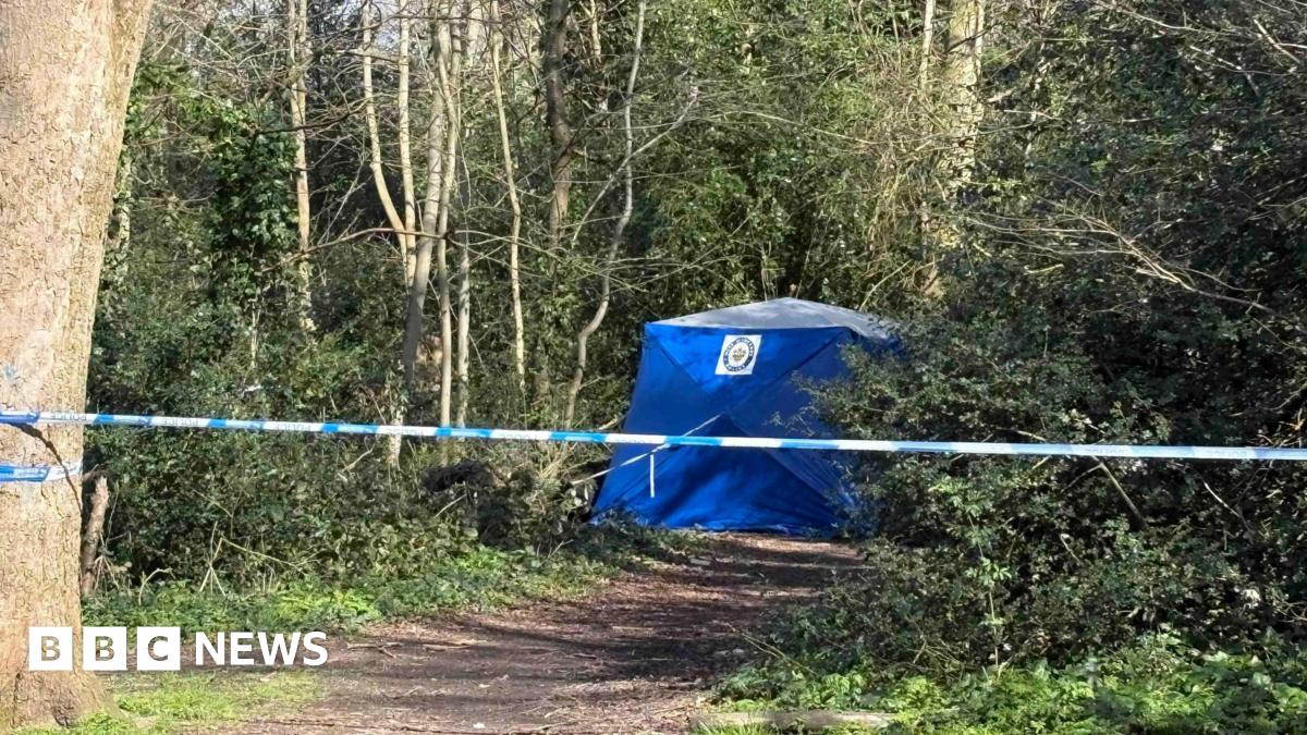 A police cordon attached to a tree in the middle of a wooded area. A blue police tent can be seen further down the footpath next to some trees.