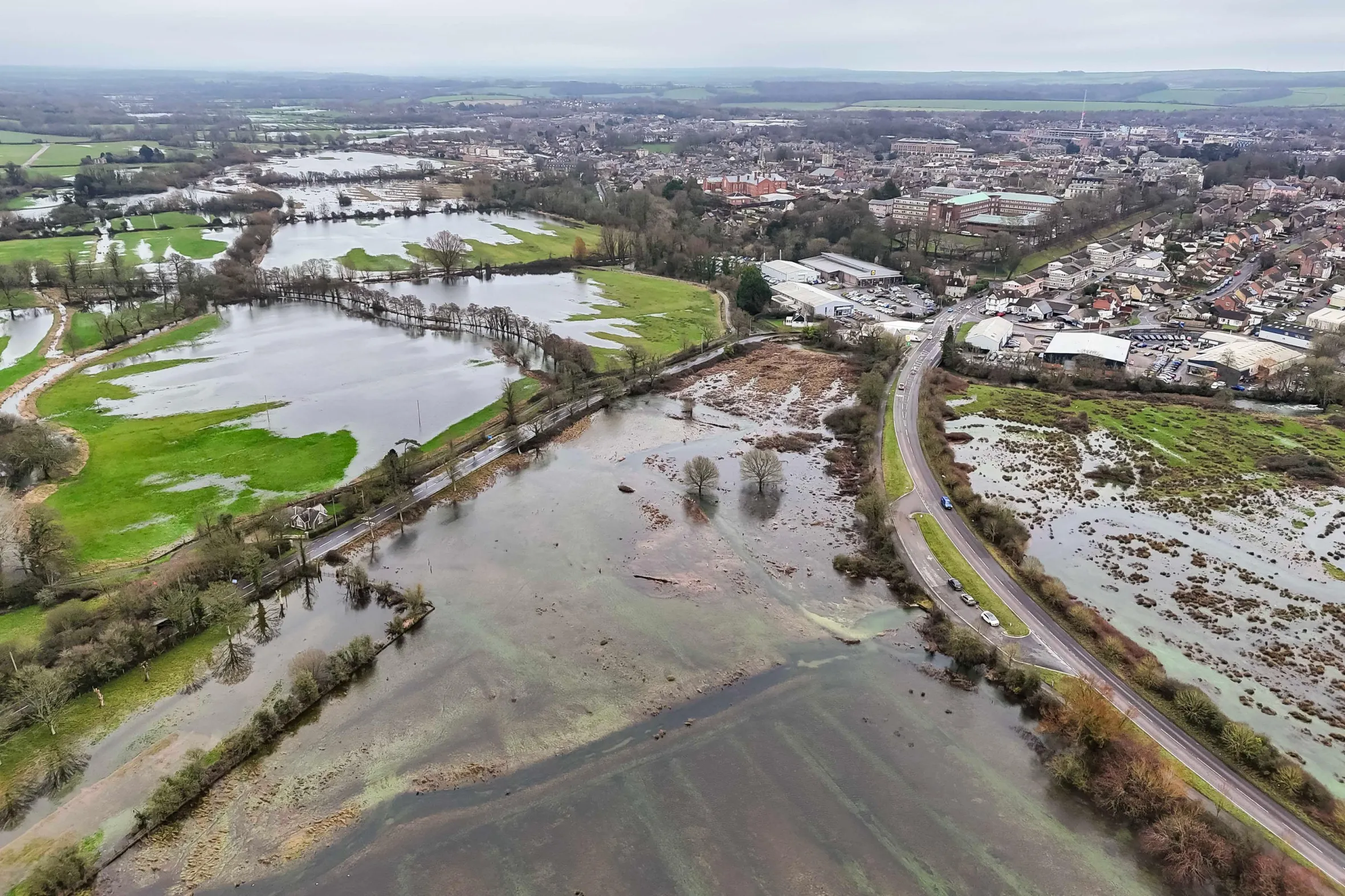 Aerial view of flooded fields in Dorchester, UK, with a road and a town visible in the distance.