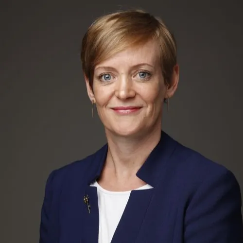 Portrait of Flora Page KC, a woman with short blonde hair and blue eyes, smiling and wearing a blue jacket with a small brooch.