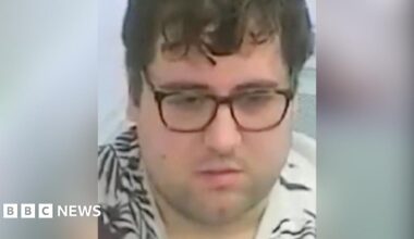 A custody image of Nathan Bennett. He has brown glasses on, a neutral expression, and mid-length brown hair. He is wearing a cream T-shirt with a brown, leaf-life design on it.