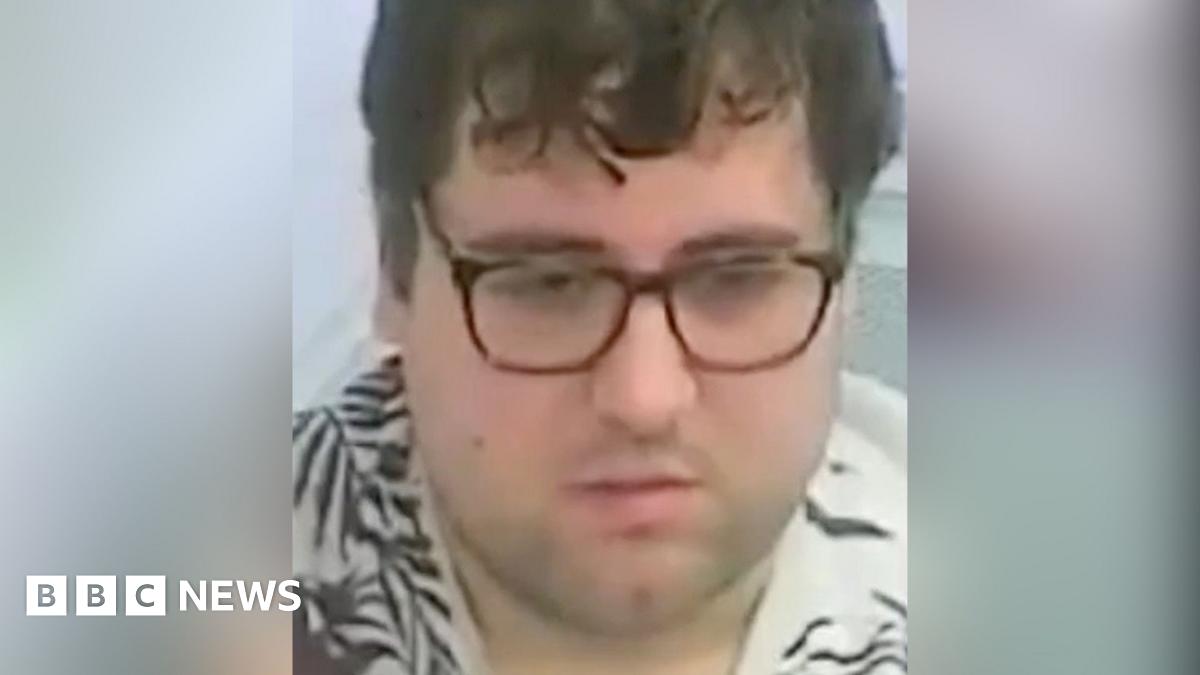 A custody image of Nathan Bennett. He has brown glasses on, a neutral expression, and mid-length brown hair. He is wearing a cream T-shirt with a brown, leaf-life design on it.