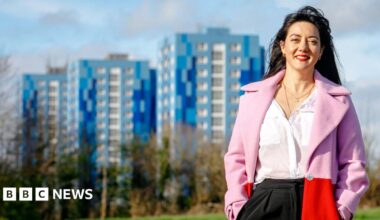 Sarah stands outdoors in a park with trees at the edges, behind which are the blue tower blocks of Marsh Farm. She wears a white blouse  under a pink and red open coat, and has long dark hair.