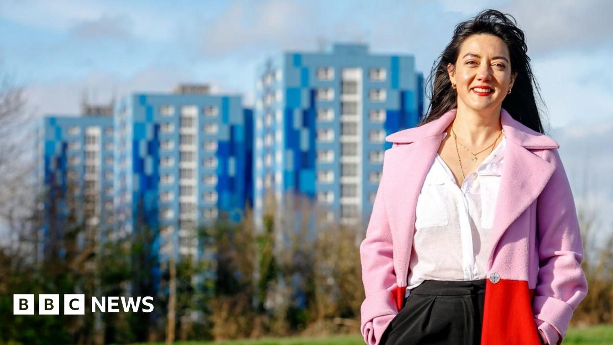 Sarah stands outdoors in a park with trees at the edges, behind which are the blue tower blocks of Marsh Farm. She wears a white blouse  under a pink and red open coat, and has long dark hair.