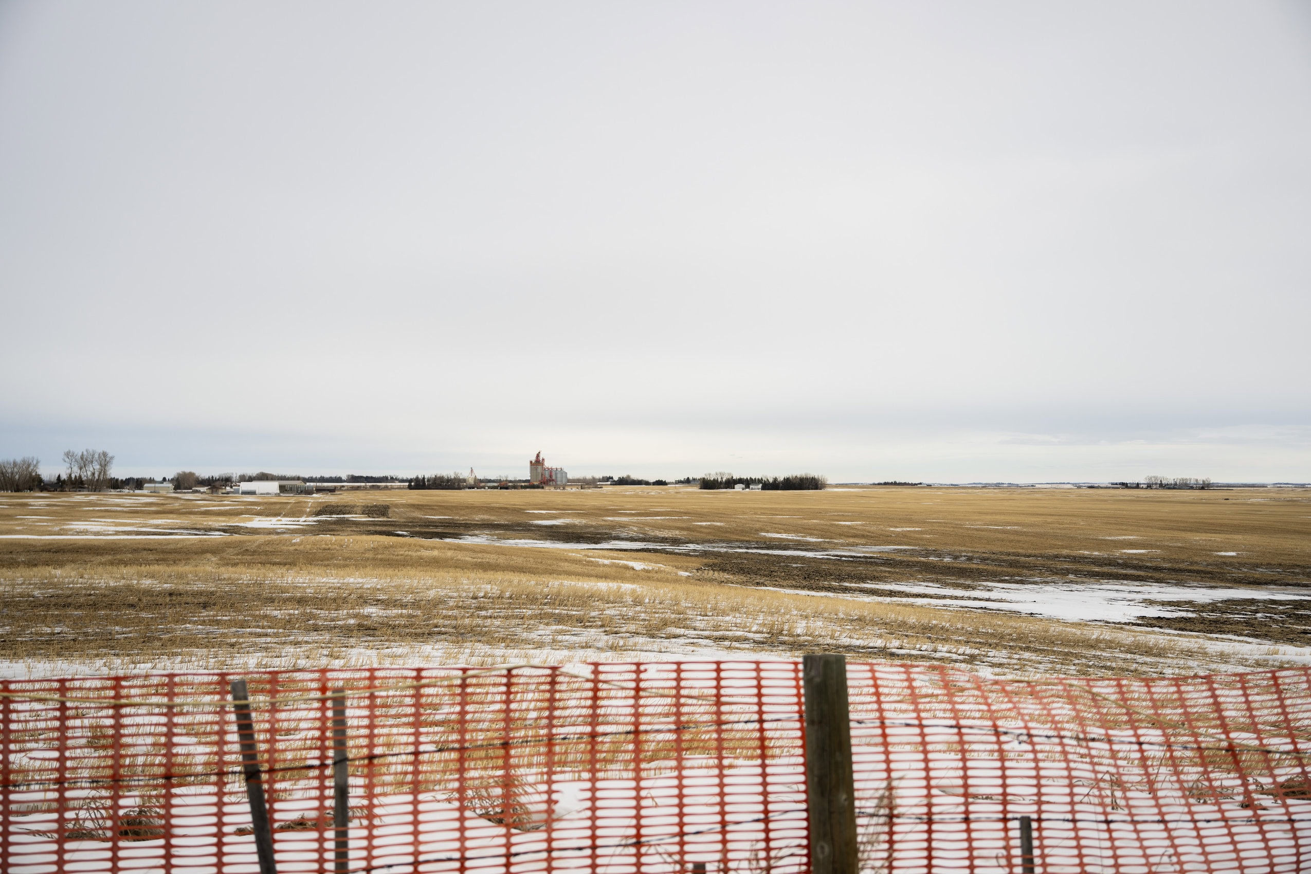 An empty field, with patches of snow and orange fencing in the foreground