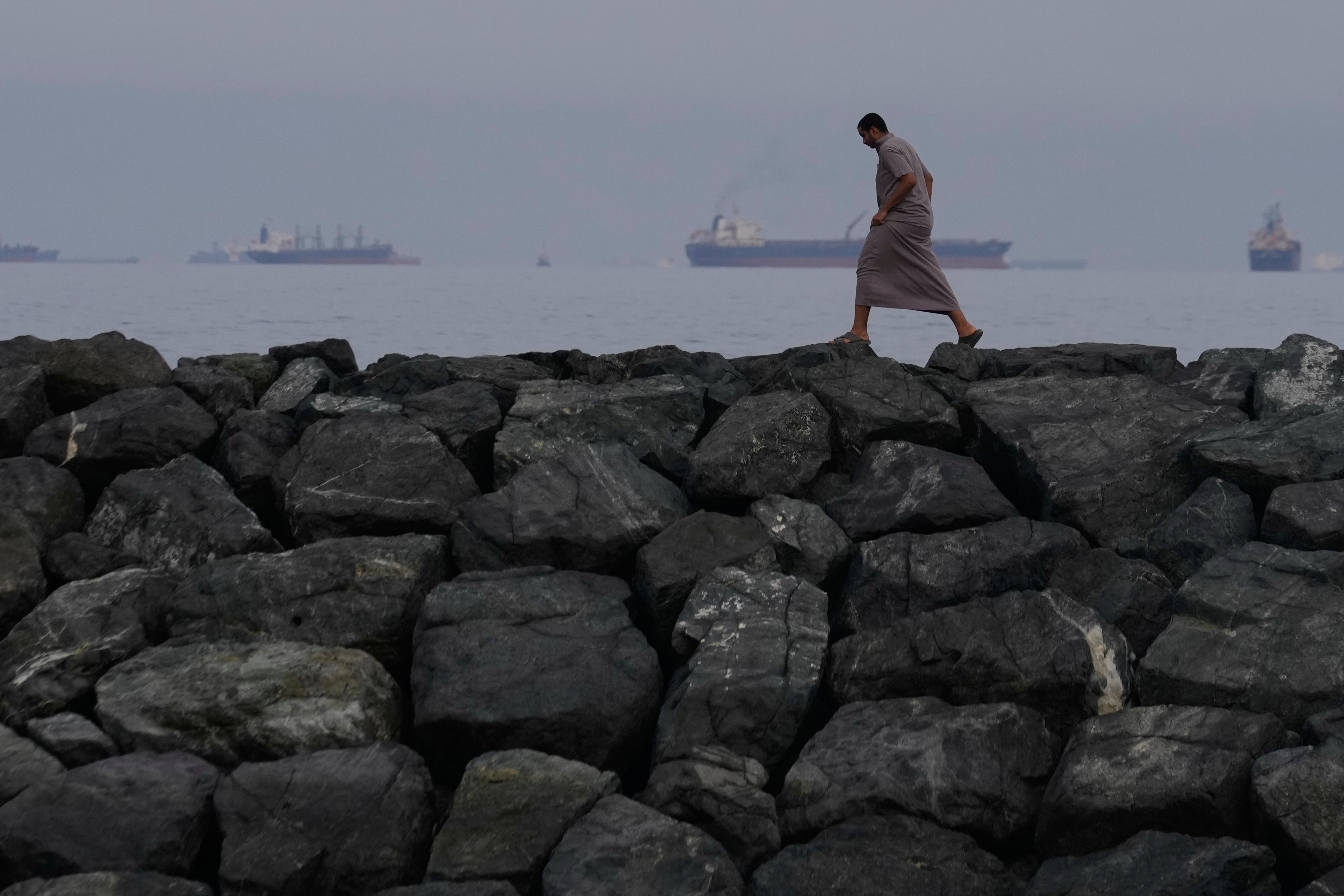 A man walks along the shore as oil tankers and cargo ships line up in the Strait of Hormuz