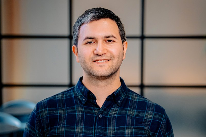 Amir Abdollahi in a professional portrait wearing a dark plaid button-down shirt against a softly blurred interior background.