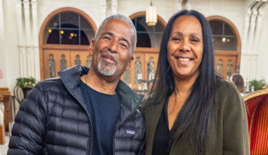 A smiling man in a black puffer jacket and jeans stands next to a smiling woman in a green coat in an ornately decorated room with arched doorways in the background.