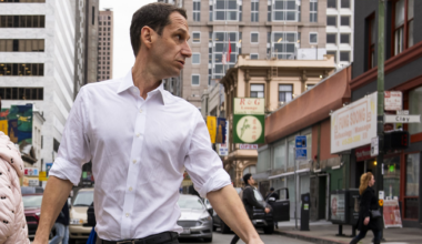 A man in a white shirt crosses a busy city street, turning his head to the side. Buildings, people walking, and cars are visible in the background.