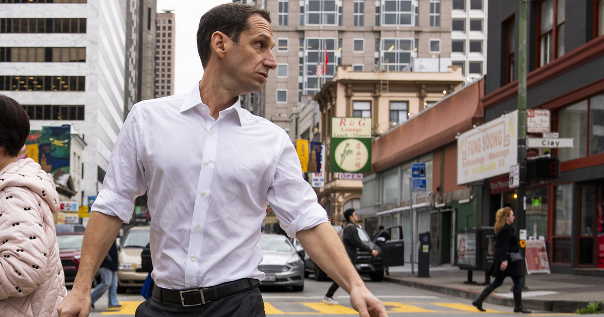 A man in a white shirt crosses a busy city street, turning his head to the side. Buildings, people walking, and cars are visible in the background.