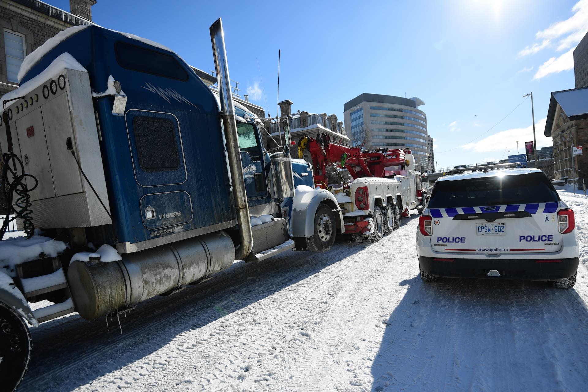 A shot of a heavy-duty tow truck pulling a blue transport truck on a snowy street, under a bright blue sky. There is a police SUV in the right foreground.