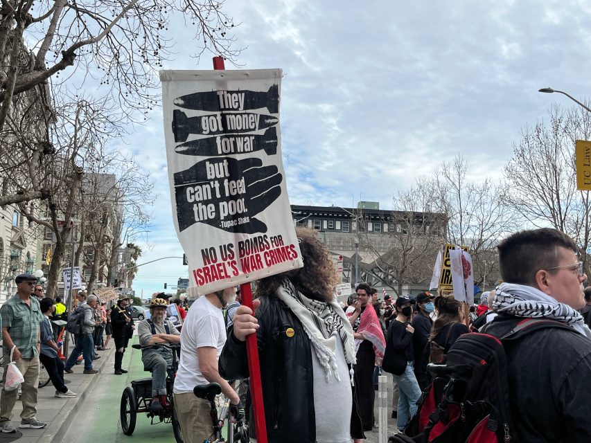 A person in a crowd holds a sign reading "They got money for war but can't feed the poor. No US bombs for Israel's war crimes" at a street protest.
