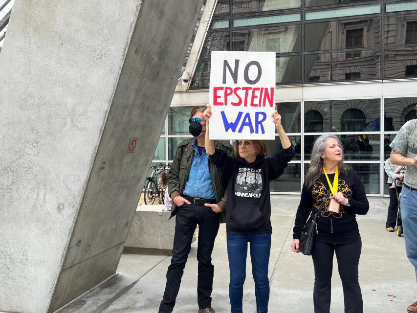 Three people stand outside a building; one holds a sign that reads "NO EPSTEIN WAR.