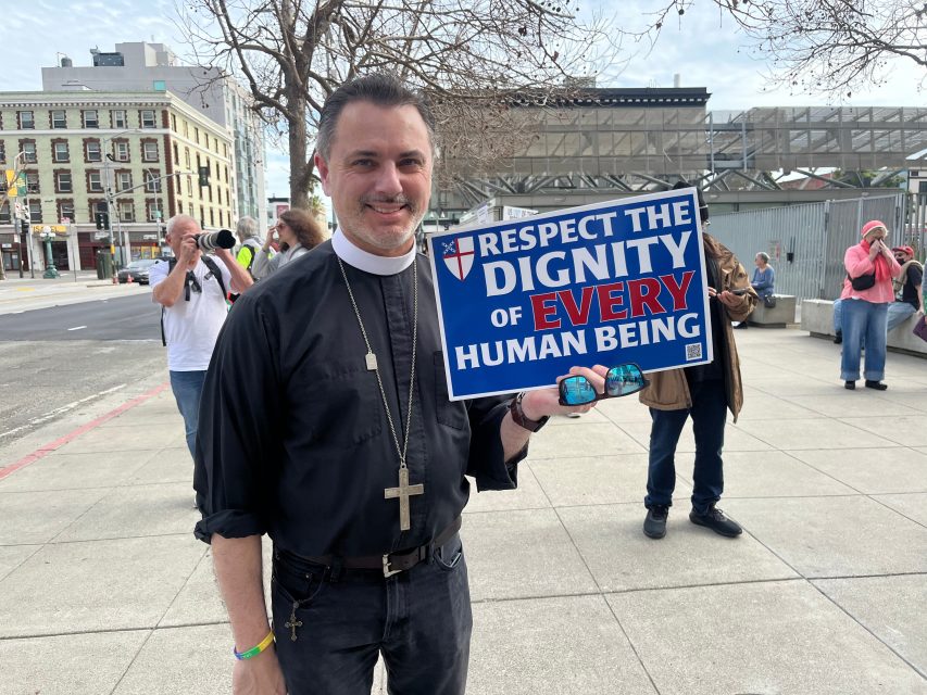 A man in clerical clothing stands outside holding a sign that reads, "Respect the dignity of every human being." Other people are visible in the background.