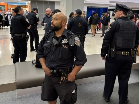 Several police officers stand and converse in a busy indoor public area, with one officer in the foreground looking to the side.