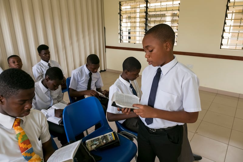 A group of young men sits in a priesthood class in Ghana. One young man is standing and reading from the scriptures.
