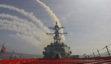 A missile launches from the deck of a U.S. Navy warship, leaving a thick trail of white smoke across a hazy sky.