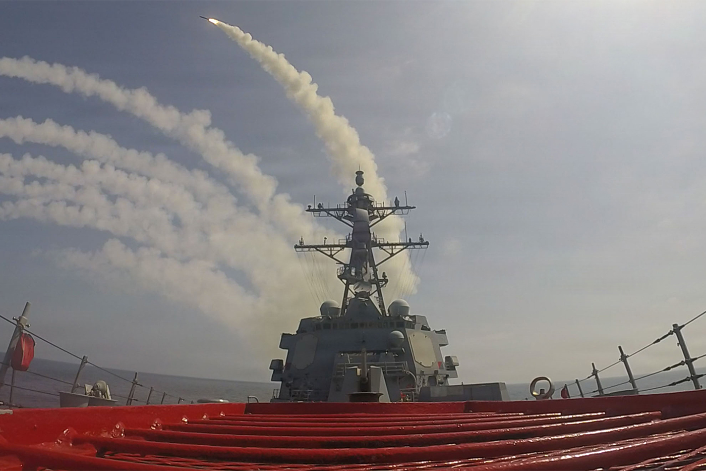 A missile launches from the deck of a U.S. Navy warship, leaving a thick trail of white smoke across a hazy sky.