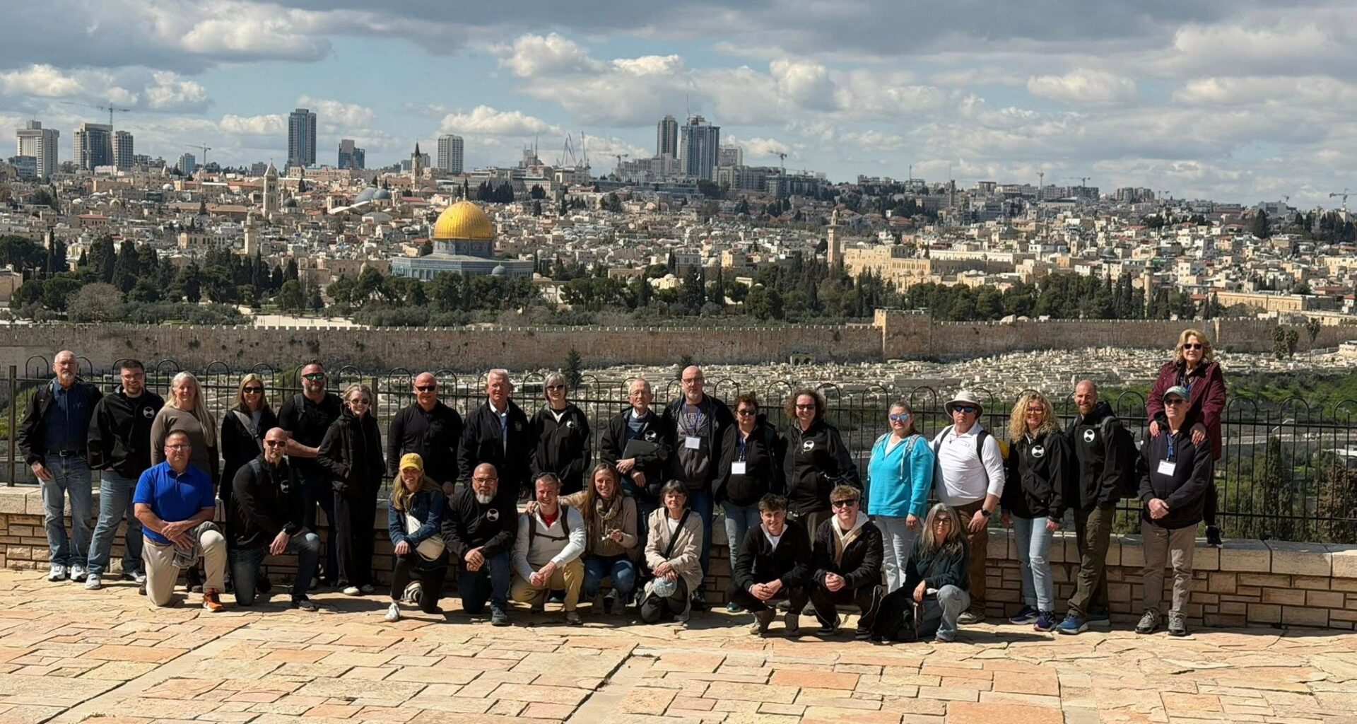 A group of people pose for a photo on a stone platform overlooking Jerusalem, with the Dome of the Rock and cityscape visible in the background under a partly cloudy sky.