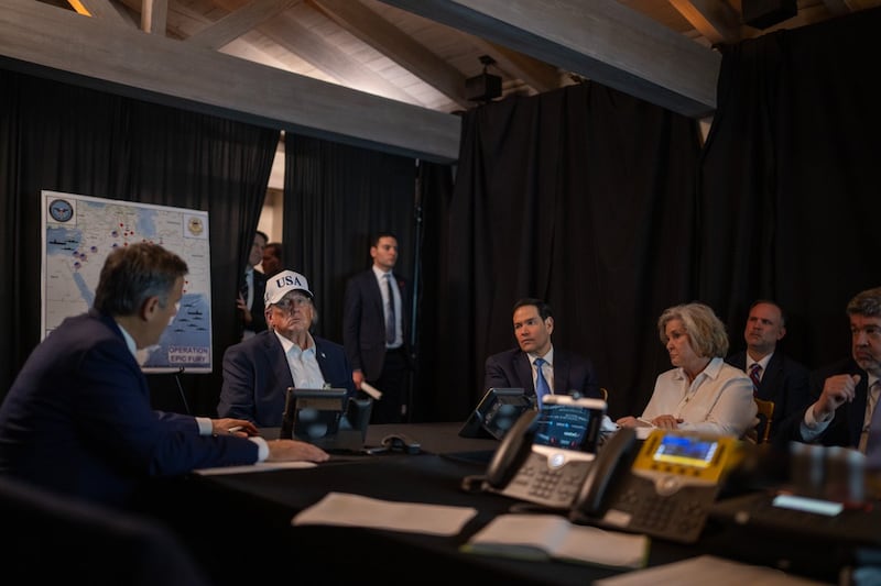 Trump in a white USA trucker hat in a darkened room with a map behind him and aides around him.