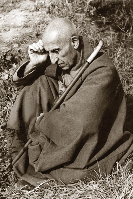 A bald man in robes sits on the ground clutching a cane.
