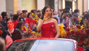 A woman in a red dress waves and smiles while sitting in a car surrounded by red and yellow flowers, with a crowd watching in the background.