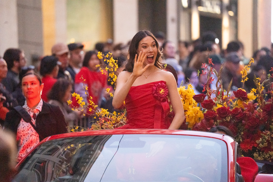 A woman in a red dress waves and smiles while sitting in a car surrounded by red and yellow flowers, with a crowd watching in the background.