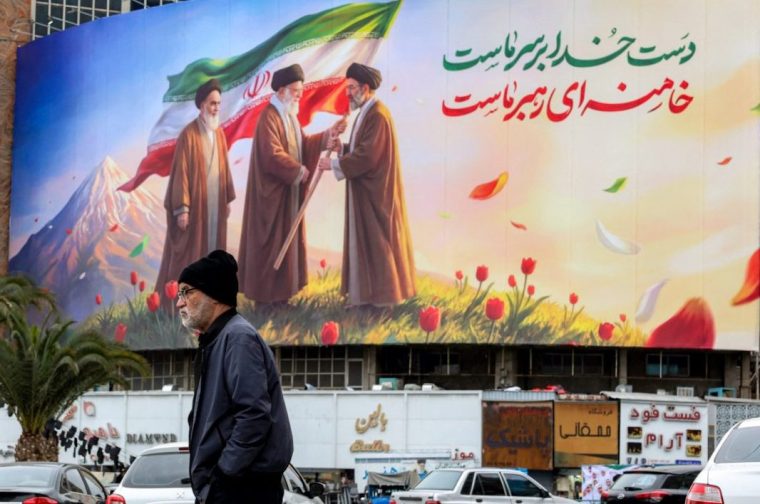 TOPSHOT - A man walks near a banner displayed at Valiasr Square in central Tehran on March 10, 2026, depicting Iran's late supreme leader Ayatollah Ruhollah Khomeini (L) watching as his successor the late Ayatollah Ali Khamenei (C) hands over a national flag to his son and new supreme leader Mojtaba Khamenei (R). Iran marked the appointment of Ayatollah Mojtaba Khamenei to replace his father as its supreme leader with a new barrage of missiles against Israel and the Gulf states on March 9, as the Middle East war sent oil prices soaring. (Photo by AFP via Getty Images) / Attention editors: AFP covers the war in the Middle East through its extensive regional network, including bureaus in Tehran, Jerusalem, and several neighboring countries. Since the start of the conflict, journalists have been working under increasingly restrictive conditions. Authorities in several countries have limited reporters' movements, photo and live video coverage from sensitive locations. Some governments and armed groups have banned images of missile or drone strikes and other security-related sites. /