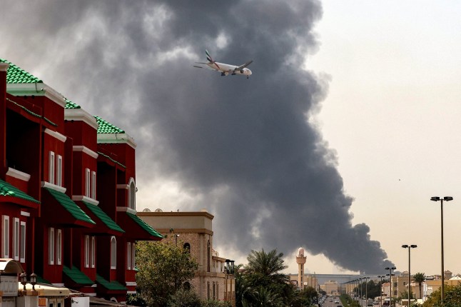 An Emirates aircraft prepares for landing as a smoke plume rises from an ongoing fire near Dubai International Airport in Dubai on March 16, 2026. Flights were gradually resuming at Dubai airport on March 16, previously the world's busiest for international flights, the airport operator said, after a "drone-related incident" sparked a fuel tank fire nearby, as Iran kept up its Gulf attacks. (Photo by AFP via Getty Images) /