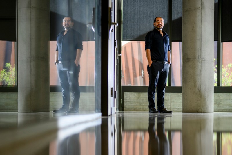 Safa Jamali stands between concrete columns in a modern building interior with reflective floors that mirror his image, natural light visible through windows in the background.