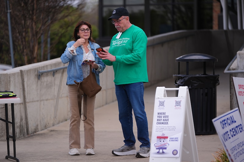 A photograph of a man and woman standing next to each other outside of a polling location looking at a cellphone.