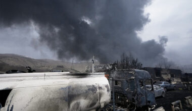 Burned industrial vehicles and equipment sit in the foreground as thick black smoke rises into the sky, with damaged buildings and hills visible in the background.