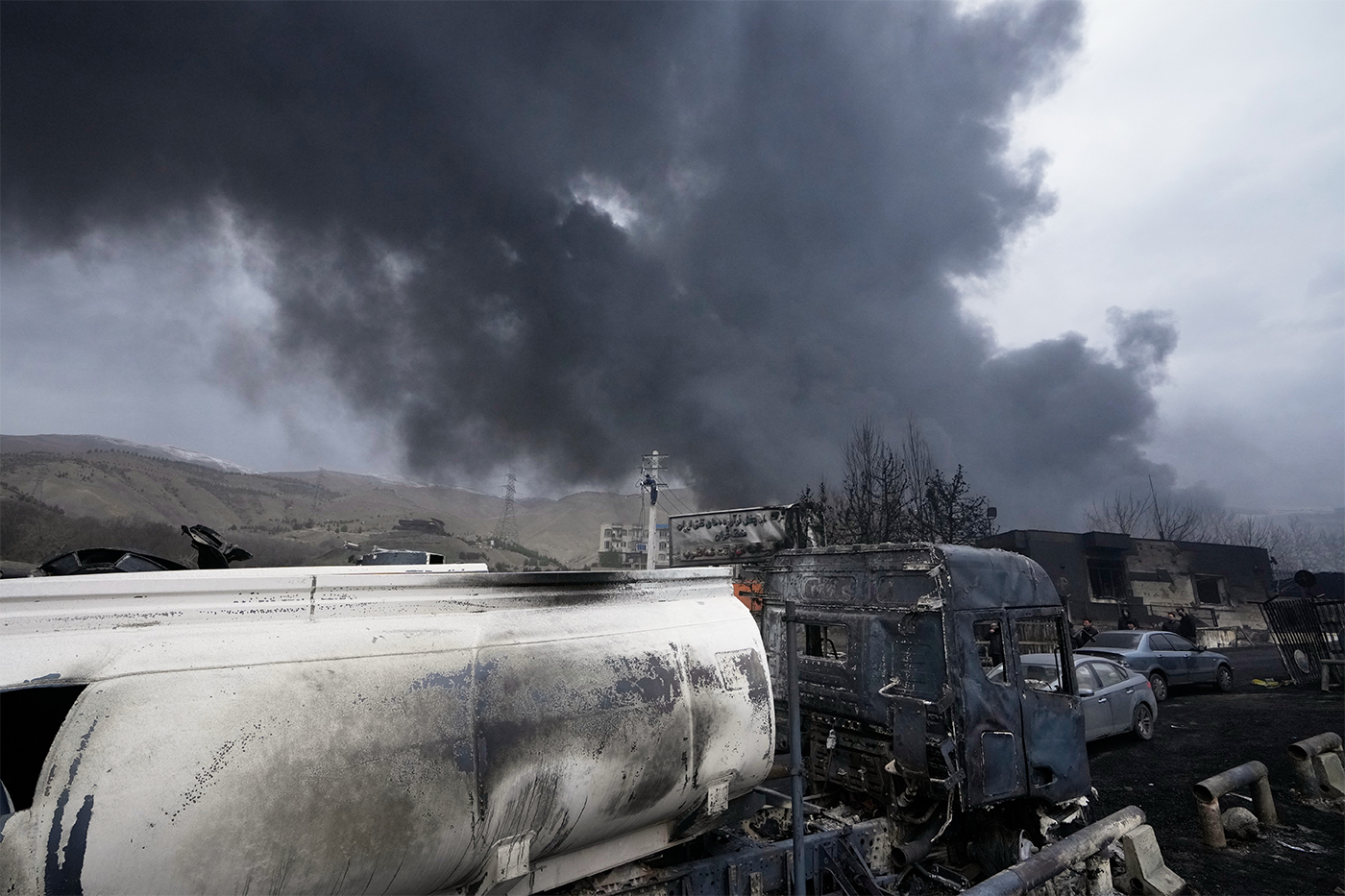Burned industrial vehicles and equipment sit in the foreground as thick black smoke rises into the sky, with damaged buildings and hills visible in the background.