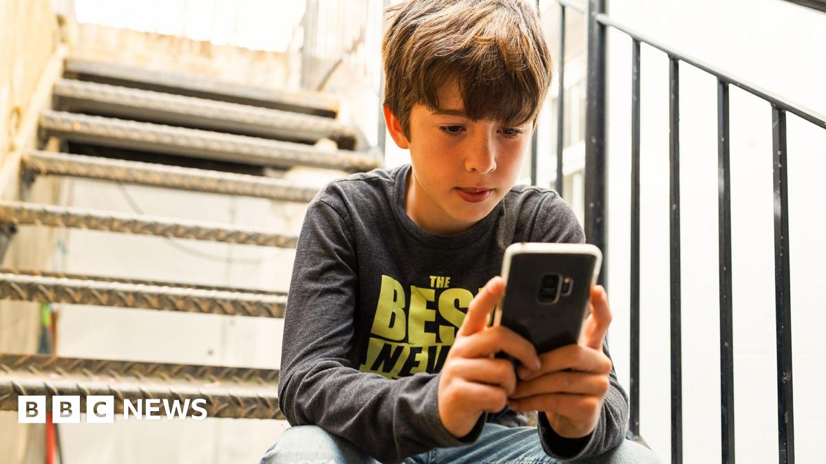 Boy aged about 14 looking at his phone. He is sitting on metal steps outside and wearing a grey t-shirt with neon yellow writing.