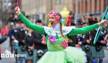A woman dressed in a green fairy outfit. She has pink flowers around her head and holding a wand in one hand and a hoola-hoop in the other.