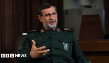 Alireza Tangsiri sits in an interview, gesturing while speaking, wearing glasses and a dark green military uniform, his greying beard and dark hair short.