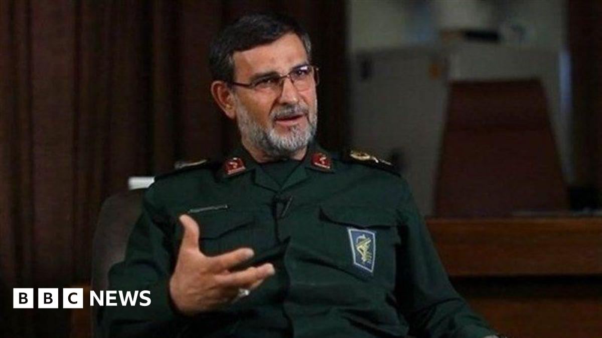 Alireza Tangsiri sits in an interview, gesturing while speaking, wearing glasses and a dark green military uniform, his greying beard and dark hair short.