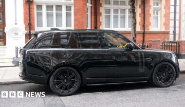 A black SUV is parked outside a brick building in London. A yellow crook lock can be seen on the wheel