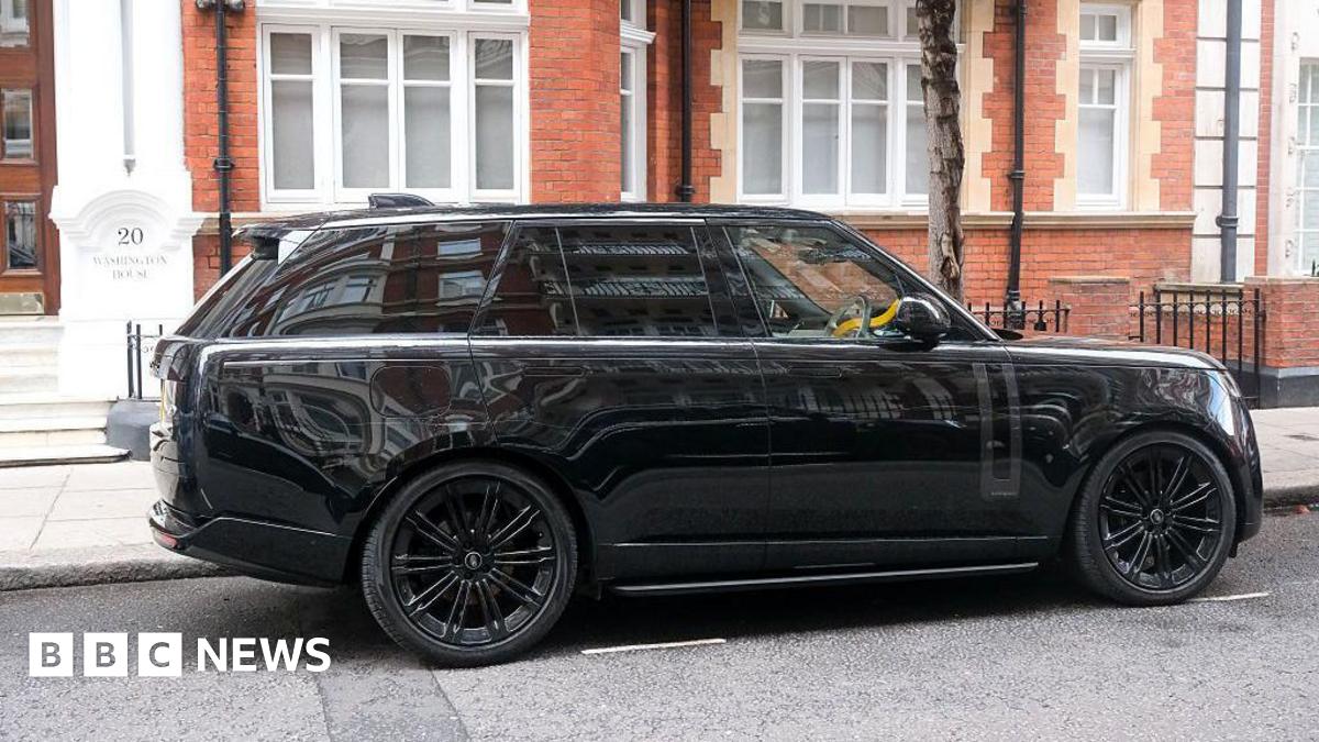 A black SUV is parked outside a brick building in London. A yellow crook lock can be seen on the wheel