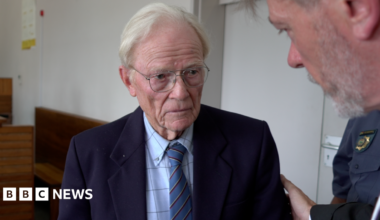 Iain Wares is wearing a navy suit jacket, blue checked shirt and blue tie. He has white hair and glasses. A man is placing a hand on his shoulder as they speak. A court of police officer can be seen in the background.