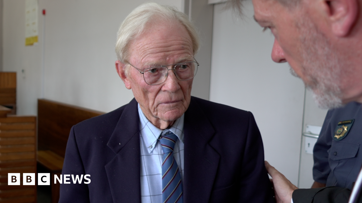 Iain Wares is wearing a navy suit jacket, blue checked shirt and blue tie. He has white hair and glasses. A man is placing a hand on his shoulder as they speak. A court of police officer can be seen in the background.