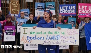 A young woman dressed in colourful headscarf and medical scrubs is holding a sign saying 'I am part of the 85% of Doctors against assisted dying'. She appears to be talking. Behind her, a group with placards supporting the proposed legislation is standing in a row. Their mouths are closed, suggesting silence.