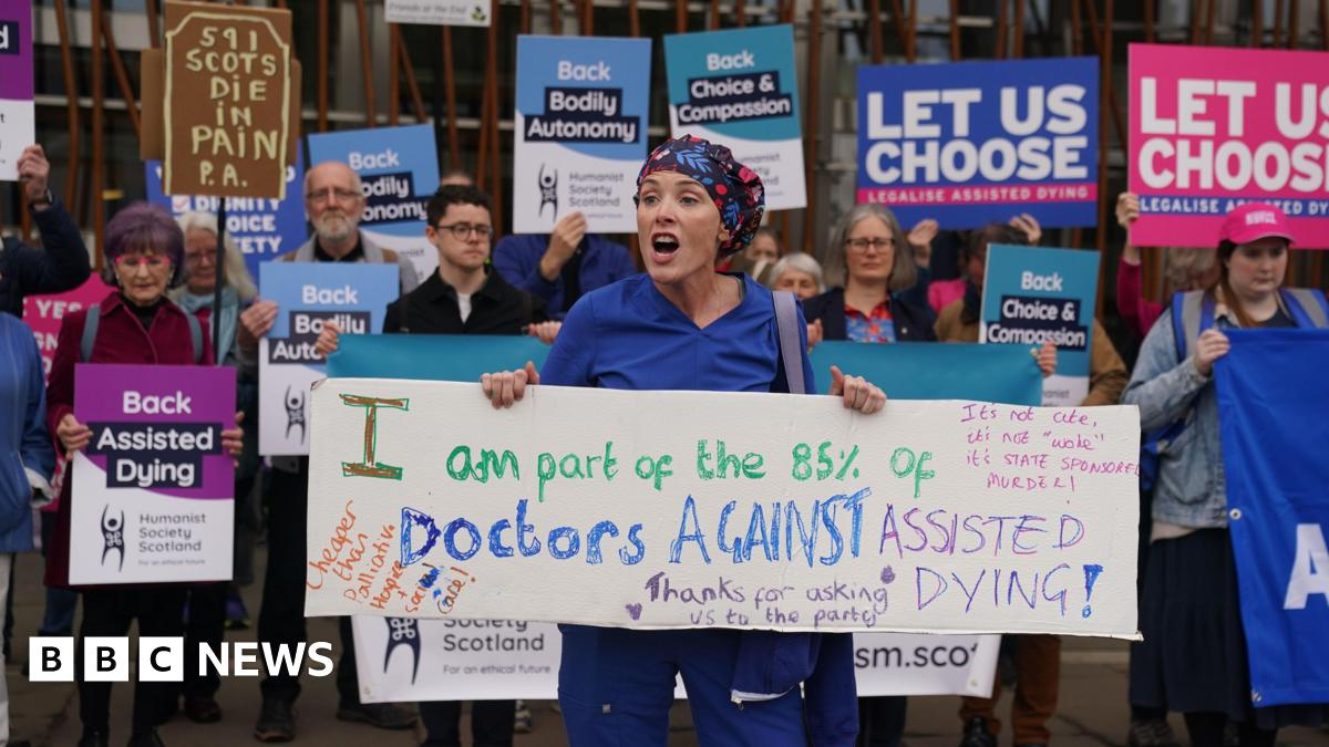 A young woman dressed in colourful headscarf and medical scrubs is holding a sign saying 'I am part of the 85% of Doctors against assisted dying'. She appears to be talking. Behind her, a group with placards supporting the proposed legislation is standing in a row. Their mouths are closed, suggesting silence.