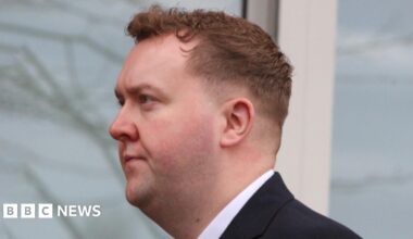 A man in a suit standing in front of a North Lanarkshire Council backdrop featuring repeated text promoting live, learn, work, invest, and visit