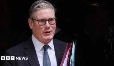 Sir Keir Starmer is wearing a dark coloured site, white shirt and blue tie. He is walking while carrying a red folder and a black folder.