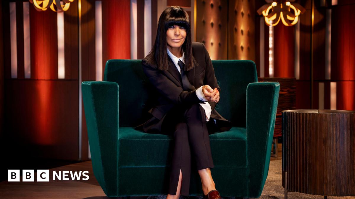 Claudia Winkleman sits on a green velvet sofa wearing a burgundy blazer and trousers