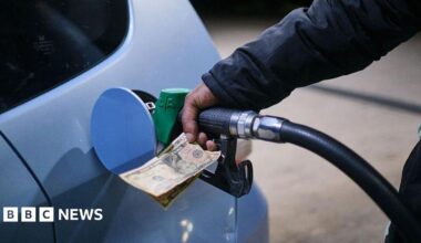 A hand holding a petrol pump into a car as it fills up at a petrol station in Zimbawe. The same hand also holds paper money. The car is silver.