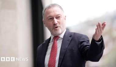 Steve Reed, a man in his late fifties with short grey hair and a white beard, speaking and looking to the right wearing a suit and red tie pointing his left arm towards the camera.