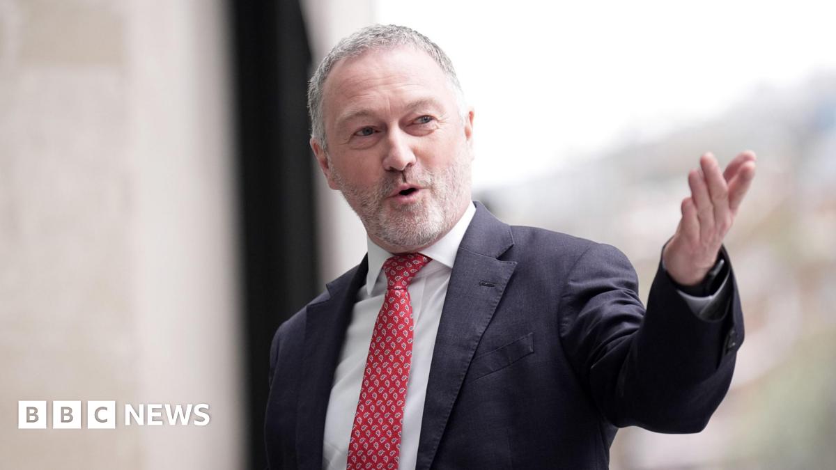 Steve Reed, a man in his late fifties with short grey hair and a white beard, speaking and looking to the right wearing a suit and red tie pointing his left arm towards the camera.