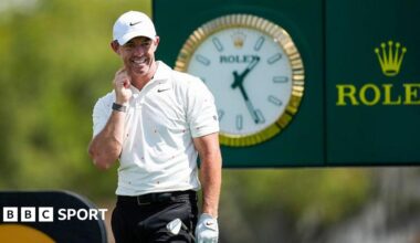 Rory McIlroy looks on after firing his tee-shot at the 14th hole of the Arnold Palmer Invitational. He is wearing a white cap, white Nike polo shirt and black trousers. He is holding his club in his gloved left hand. A large Rolex clock and advertising board can be seen behind him.